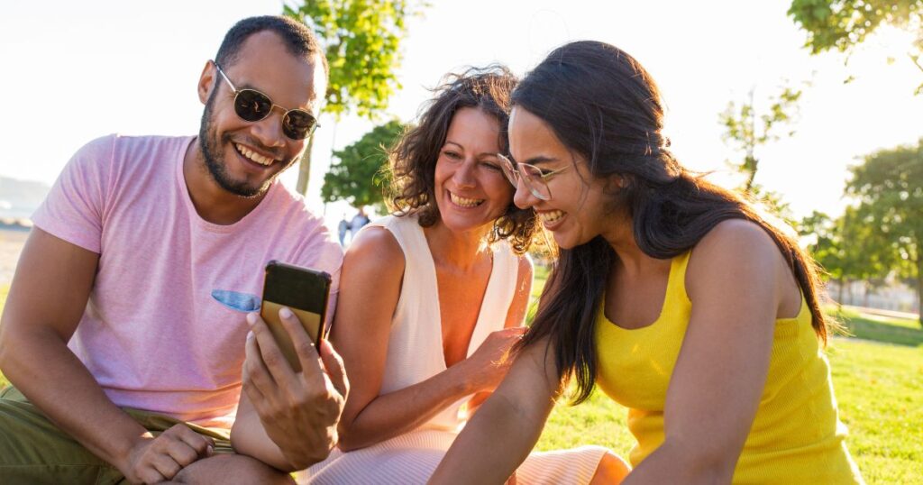 Happy Latin guy sharing funny news with his female friends. Man and women sitting on grass in park, looking at smartphone screen and laughing. Content on phone concept